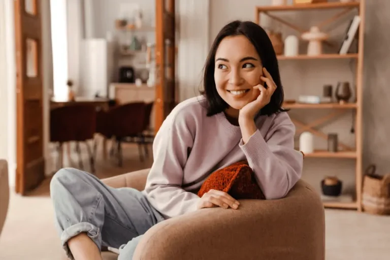 Smiling young woman sitting comfortably in a cozy living room, representing happiness, confidence, and a healthy lifestyle.